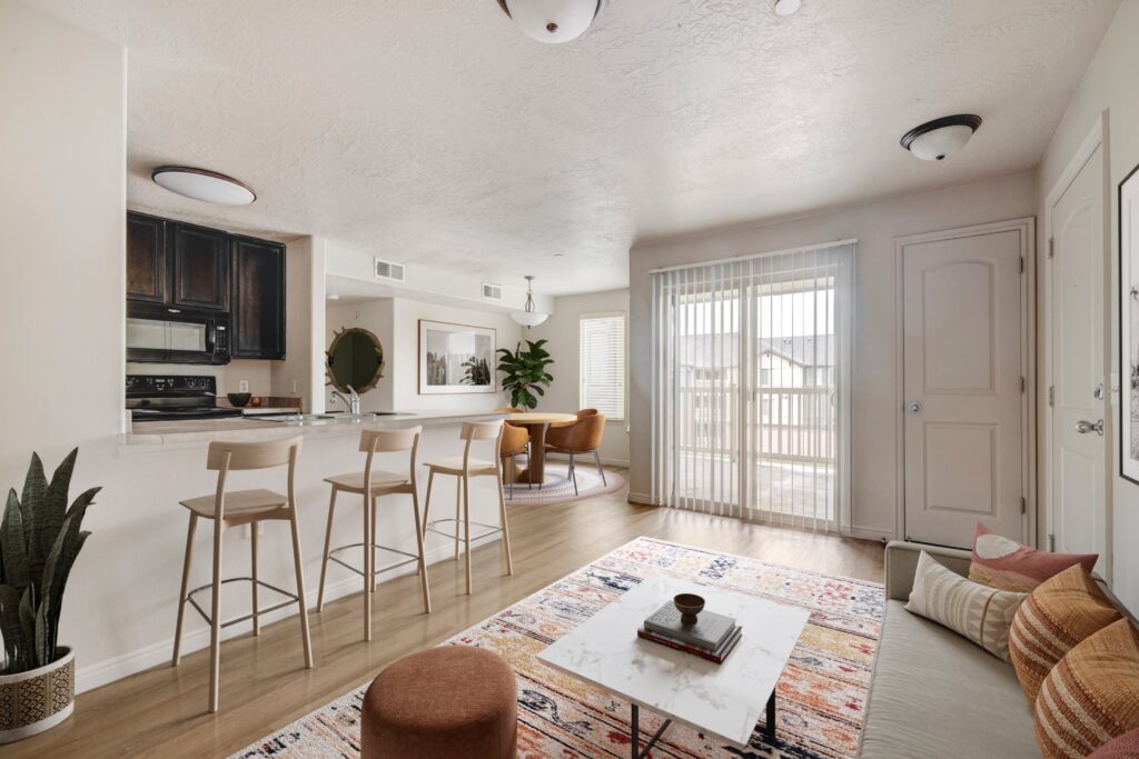 An open-concept living space with a kitchen breakfast bar, wooden barstools, a round dining table with cushioned chairs, a patterned area rug, a white coffee table, and a sofa with decorative pillows.