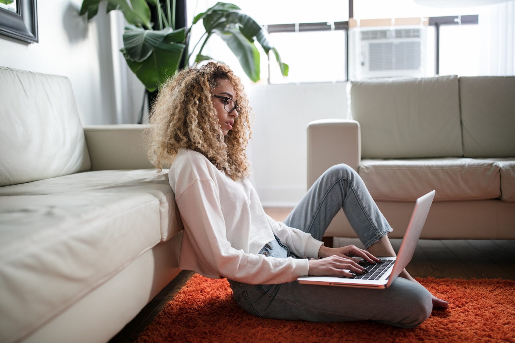 A person with curly hair and glasses sits on an orange rug, leaning against a light-colored sofa while using a laptop. Large windows and green plants are visible in the background, creating a bright, cozy atmosphere.