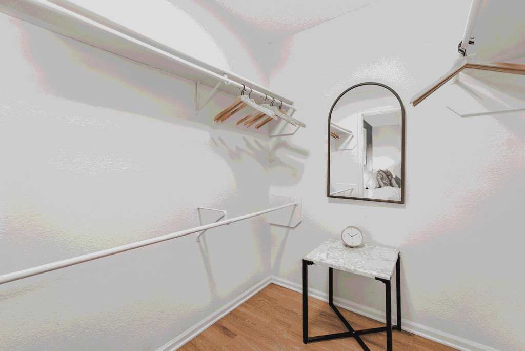 Minimalist walk-in closet with white walls, empty white shelves, wooden hangers, a small marble-top table with a clock, and an arched mirror reflecting part of a bed. The floor is light wood.