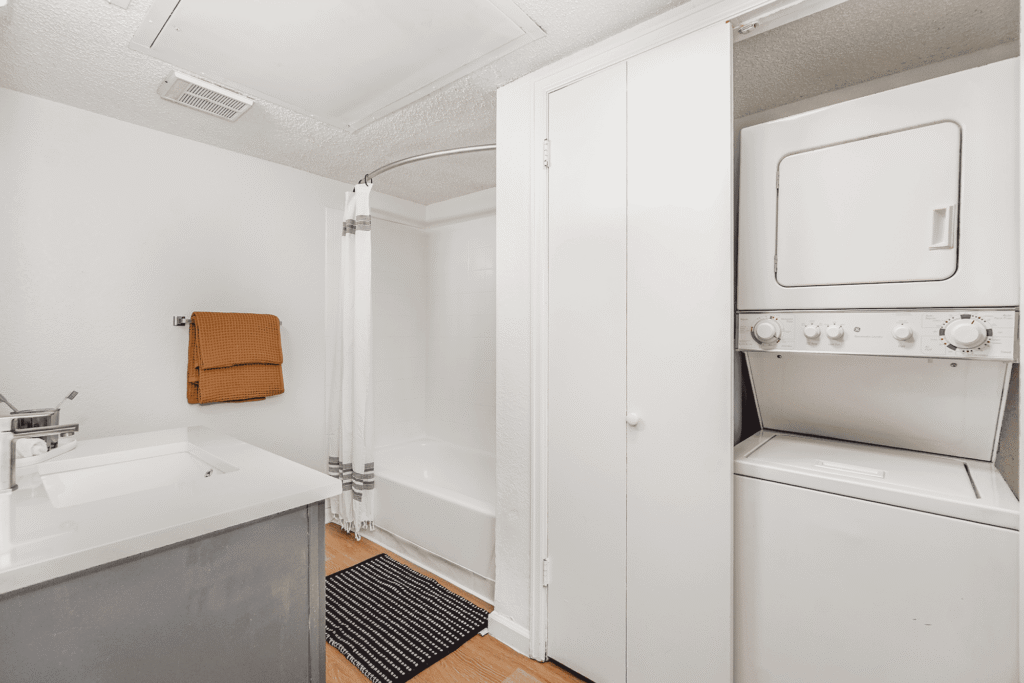 Modern bathroom with white walls, a bathtub and shower, a stacked washer and dryer in a closet, a sink with a gray vanity, a towel on the wall, and a black-and-white rug on a wooden floor.