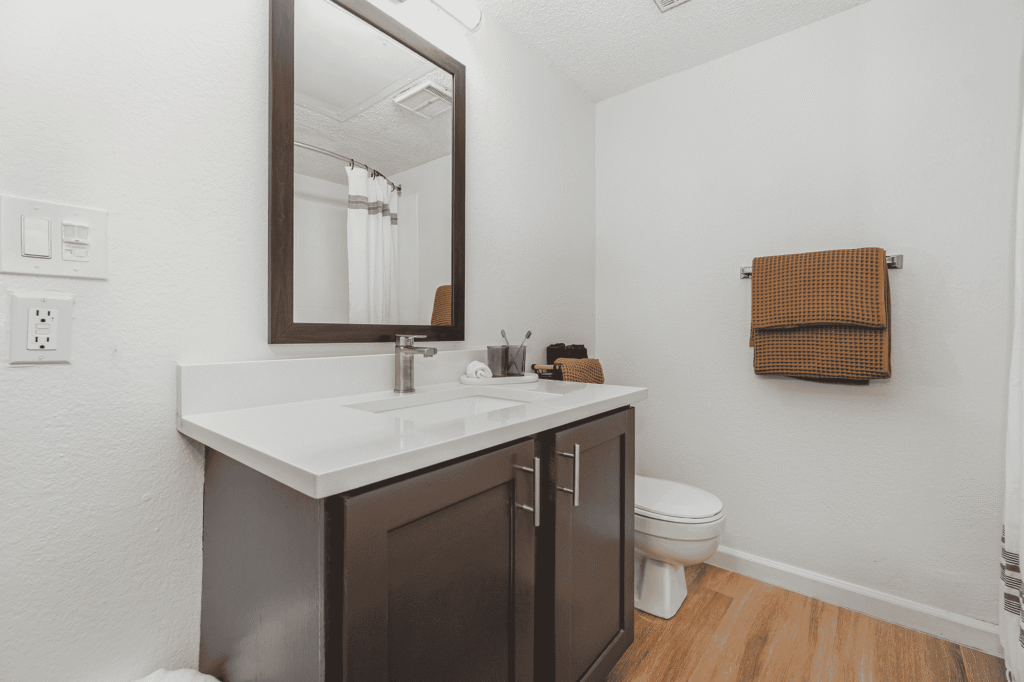 Modern bathroom with a dark wood vanity, large mirror, white countertop, and faucet. Brown towels hang on a wall rack. The floor is wood, and the walls are plain white. A toilet sits next to the vanity.
