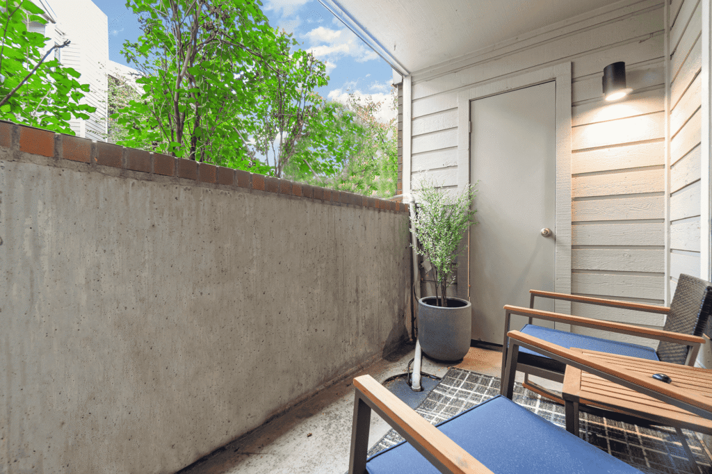 A small outdoor patio with two blue cushioned chairs, a potted plant, a wall-mounted light, and partial brick and concrete walls. Green trees are visible behind the patio wall under a blue sky.