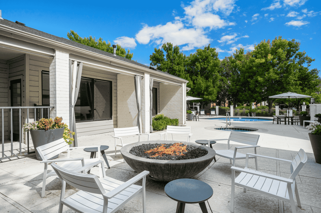 Modern outdoor patio with white chairs arranged around a fire pit, near a pool and hot tub. Trees and blue sky are in the background, and the area has a relaxed, inviting atmosphere.