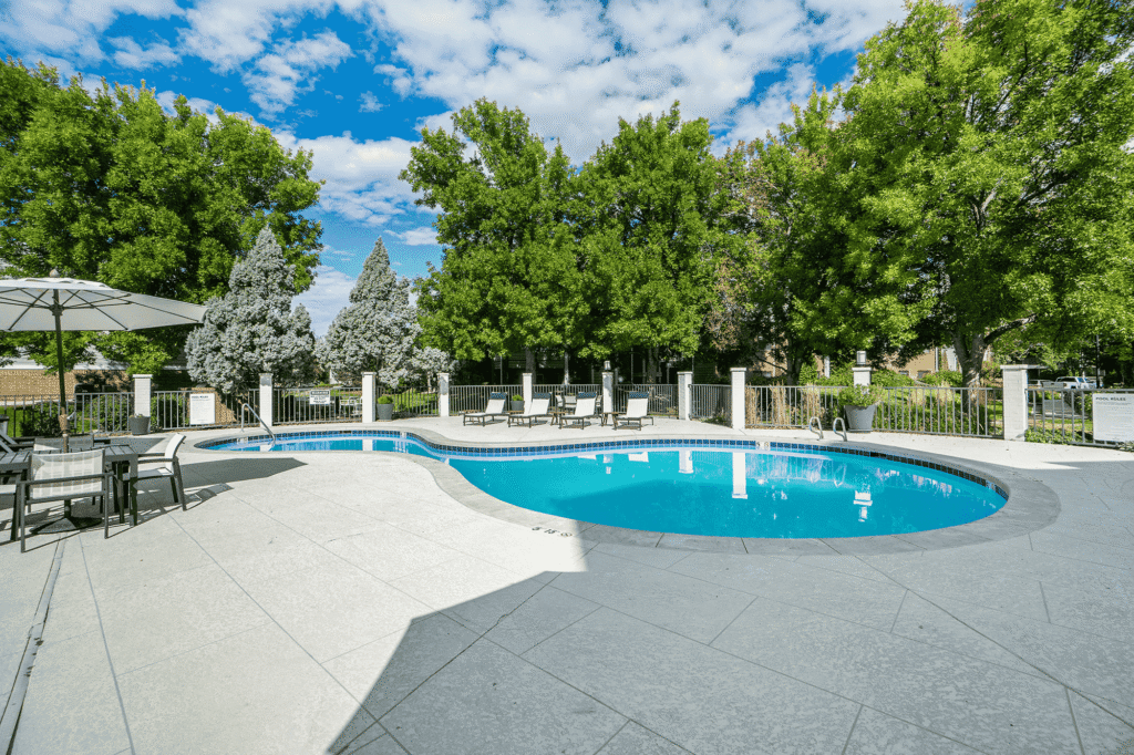 A small outdoor pool with lounge chairs, tables, and umbrellas, surrounded by a fence and lush green trees under a partly cloudy blue sky.