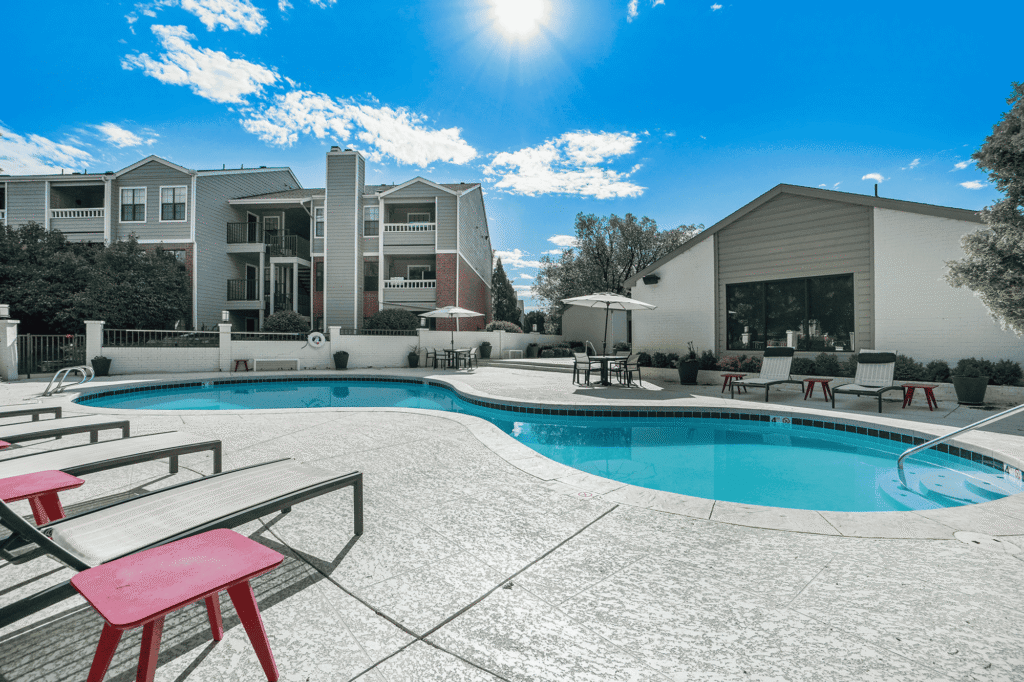 Sunny outdoor pool area with lounge chairs, red side tables, umbrellas, and nearby apartment buildings. Clear blue sky and bright sunlight create a vibrant, inviting atmosphere.