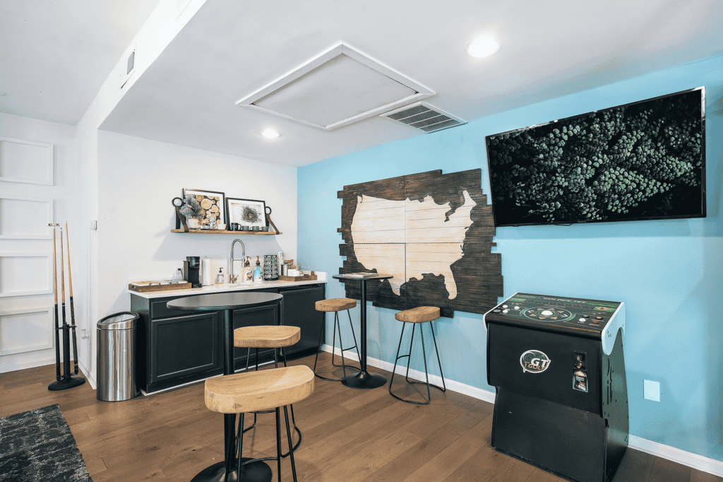 Modern game room with a blue accent wall, wall-mounted TV, arcade machine, wooden stools, small bar area with a sink, art on shelves, and pool cues in the corner. Light wood floor and white ceiling lights are visible.