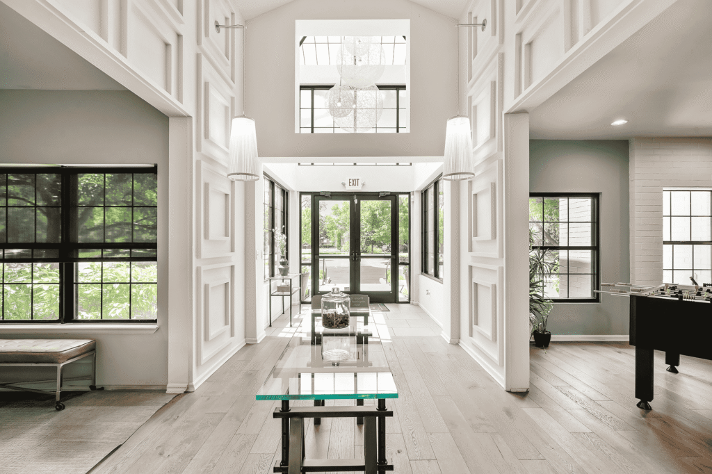 Modern, bright entryway with high ceilings, glass doors, large windows, wooden floors, white walls, a foosball table, plants, and contemporary decor, letting in abundant natural light.