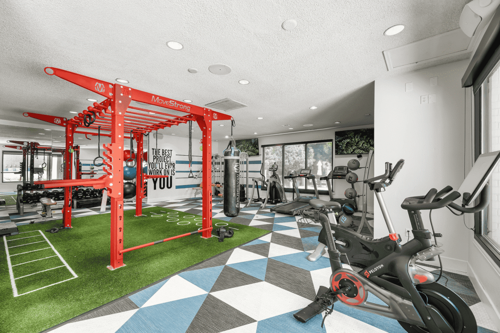 Modern gym with red workout equipment, stationary bikes, treadmills, punching bag, squat rack, turf area, and geometric blue and gray flooring. Motivational quote on the wall: The best project youll ever work on is you..