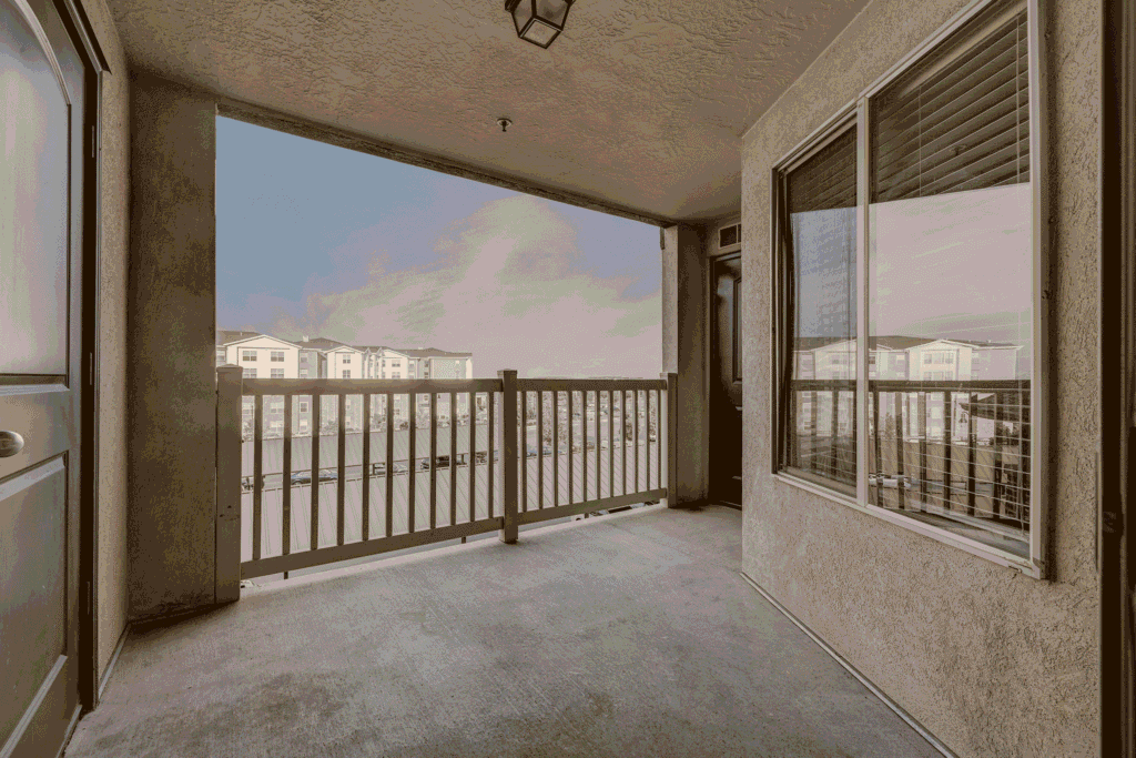 Covered apartment balcony with beige walls, railing, and two doors. Large window with blinds reflects nearby buildings. Blue sky and other apartment buildings are visible in the background.
