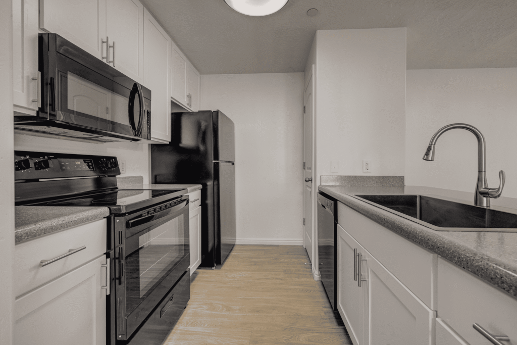Modern kitchen with white cabinets, black appliances including stove, microwave, dishwasher, and refrigerator, and a large sink with a curved faucet on a gray countertop. Light wood flooring is visible.