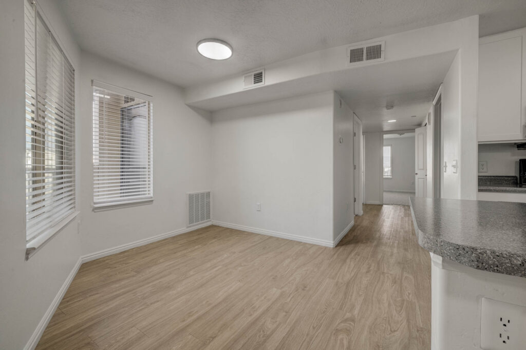 A bright, empty room with light wood floors, white walls, two large windows with blinds, and an open kitchen area visible in the background. Recessed lighting and air vents are on the ceiling.