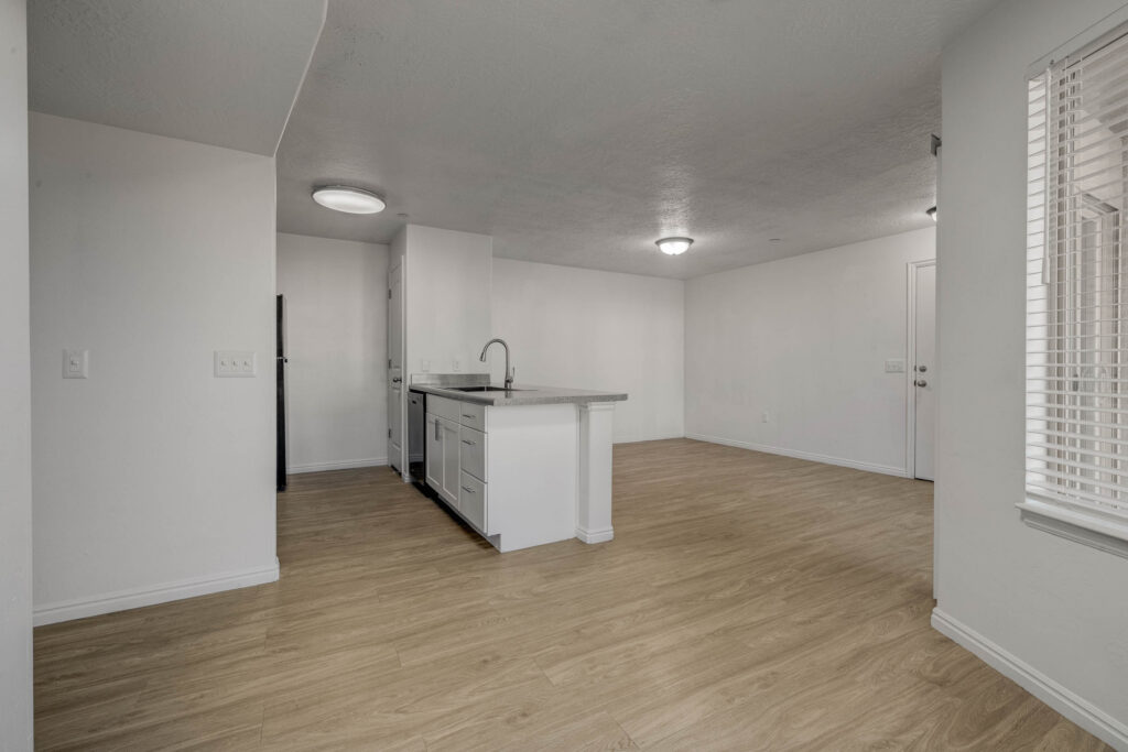 Modern apartment interior with light wood flooring, white walls, an open kitchen with an island and sink, visible appliances, and a well-lit living space with large windows and a door in the background.