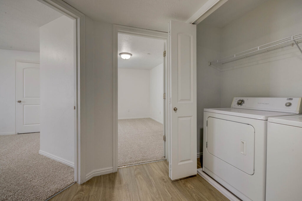 A small laundry area with a white washing machine and dryer, adjacent to a hallway with wood flooring and two carpeted rooms visible through open doors. Walls and doors are white.