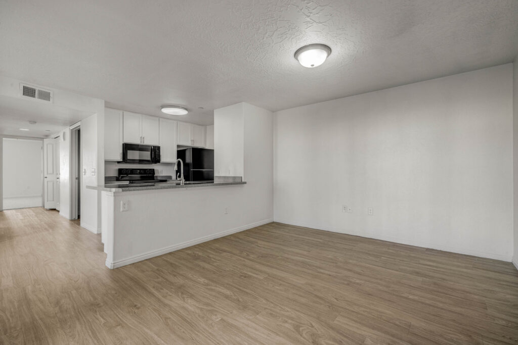 Empty apartment interior with light wood floors, white walls, and a small open kitchen featuring stainless steel appliances and gray countertops. Bright ceiling lights illuminate the space. Hallway is visible to the left.
