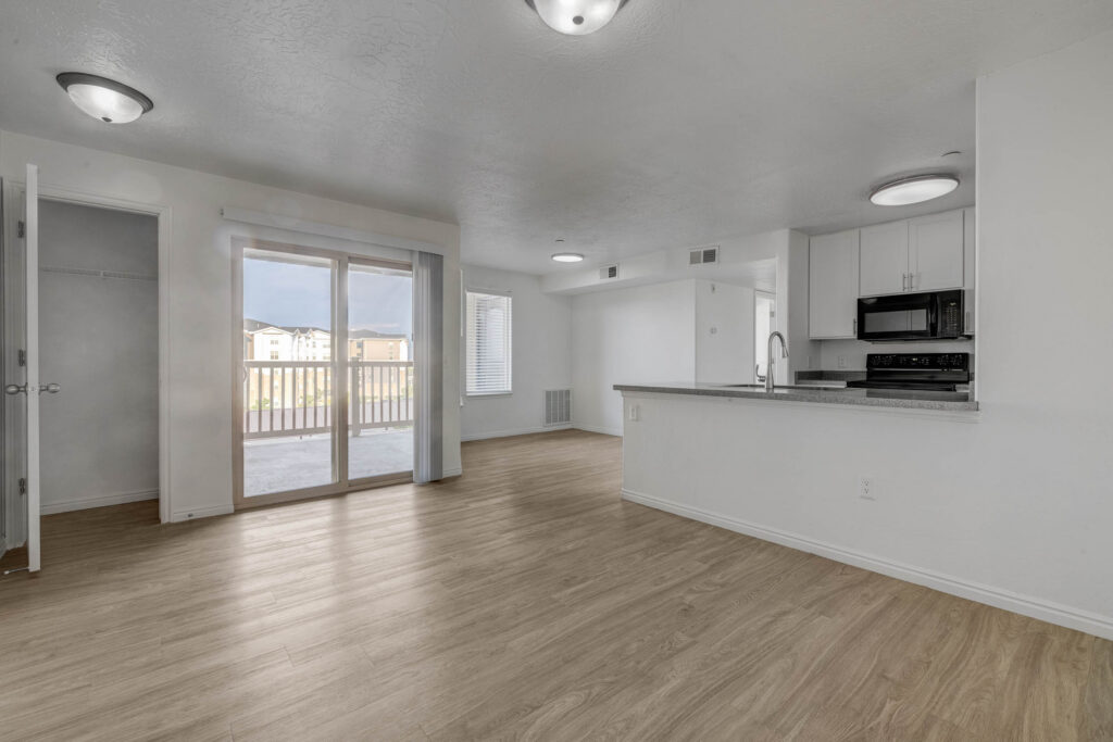 Open living area with light wood flooring, white walls, sliding glass doors to a balcony, a small closet, and a modern kitchen with white countertops and black appliances. Ceiling lights brighten the space.